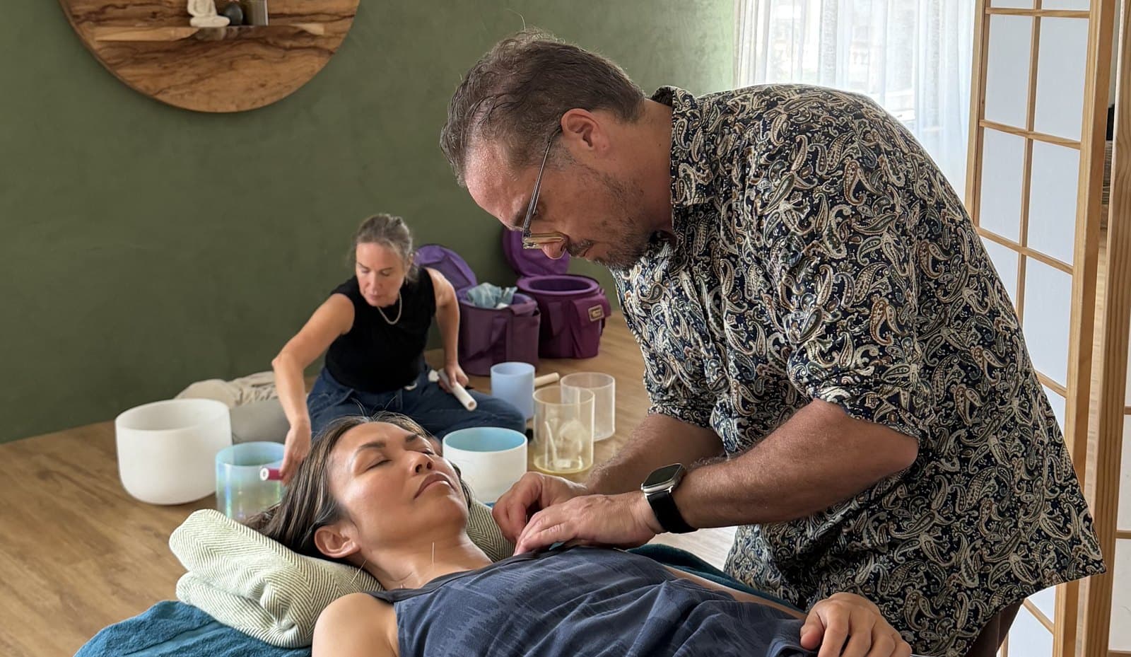 Steven working at the treatment table while Renata plays crystal singing bowls behind him at Open Sky Wellbeing